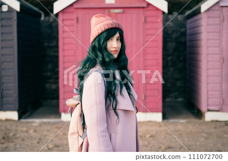 Stylish hipster woman with color hair in pink outfit and backpack walking along wooden beach huts on seaside. Off season Travel concept. Seasonal street fashion. Barbiecore style. Simple pleasures. Stylish hipster woman with color hair in pink outfit and backpack walking along wooden beach huts on seaside. Off season Travel concept. Seasonal street fashion. Barbiecore style. Simple pleasures. 111072700