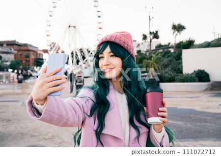 Stylish young smiling hipster woman with color hair holding reusable coffee cup wearing pink coat, knitted hat taking a selfie photo on phone on street walk, happy mood, seasonal fashion style trend Stylish young smiling hipster woman with color hair holding reusable coffee cup wearing pink coat, knitted hat taking a selfie photo on phone on street walk, happy mood, seasonal fashion style trend 111072712