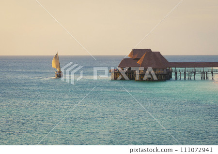 Dhow boat sails in the ocean near beautiful thatch stilt house restaurant at sunset Dhow boat sails in the ocean near beautiful thatch stilt house restaurant at sunset 111072941