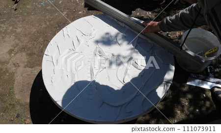 the master applies a thick layer of elastic white mastic to the round surface of the product using a long flat bar, leveling the texture of the solution, working outdoors, view from the top angle 111073911