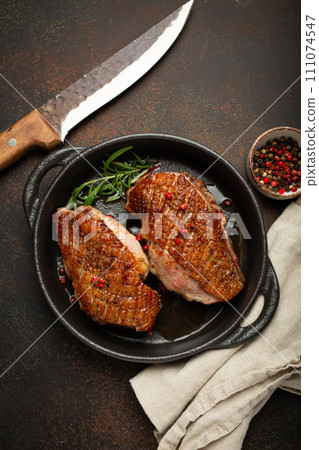 Two roasted duck breast fillets with crispy skin, with pepper and rosemary, top view in black cast iron pan with knife, dark brown concrete rustic background 111074547