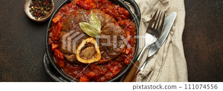 Traditional Italian dish Ossobuco all Milanese made with cut veal shank meat with vegetable tomato sauce served in black casserole pan top view on rustic brown background Traditional Italian dish Ossobuco all Milanese made with cut veal shank meat with vegetable tomato sauce served in black casserole pan top view on rustic brown background 111074556
