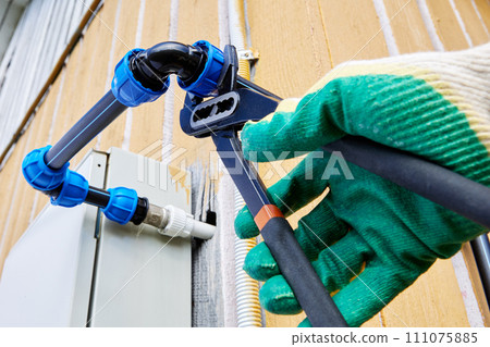 Installation of drinking and irrigation external water supply from HDPE pipes, plumber tightens fastening nut of compression clamp of elbow fitting using plumbing pliers. 111075885