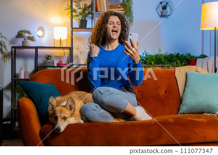 Surprised woman browsing smartphone celebrating success winning lottery sitting beside corgi dog 111077347