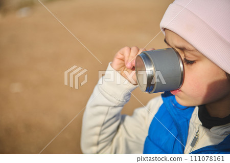 Face portrait little child girl in pink hat and blue down jacket, drinking hot tea from steel thermos mug. Copy ad space 111078161
