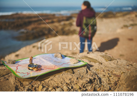 Selective focus on compass and map on the rock over blurred background of a backpack female tourist enjoying his journey Selective focus on compass and map on the rock over blurred background of a backpack female tourist enjoying his journey 111078196
