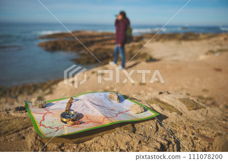 Details on a compass and map on a rock against blurred female tourist looking into the distance through binoculars 111078200