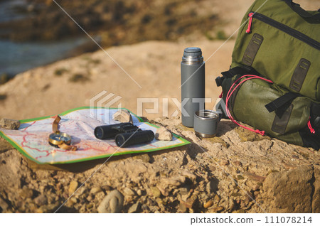 Still life backpack and thermos flask on the cliff against Atlantic ocean background while waves break on the headland. 111078214