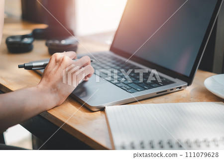 Close-up view of a person's hand holding a pen, ready to write in a notebook beside an open laptop, highlighting multitasking and productivity. 111079628