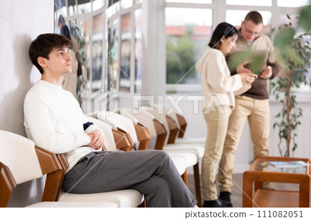Young man browsing websites on smartphone while sitting in lobby of job center or employment office, waiting for interview or conversation with career counselor 111082051