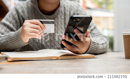 A cropped shot of a woman holding her smartphone and a credit card while sitting at a table. 111082330