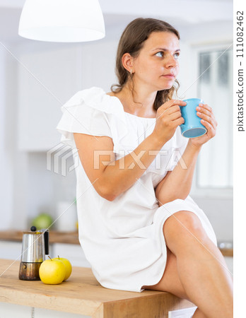 Woman sitting on kitchen countertop, drinking coffee Woman sitting on kitchen countertop, drinking coffee 111082462