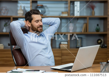 Relaxed brunette man leaning back and holding hands behind head in personal workspace with pc. Calm head of company taking break during busy day and enjoying daydreaming in comfortable chair. 111087513