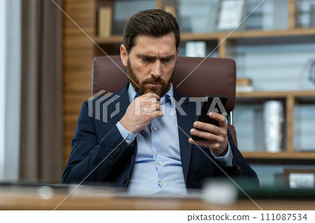 Pensive bearded guy in shirt and jacket holding cell phone and rubbing chin with worried facial expression. Concerned man reading unpleasant message and thinking about problem solution in cabinet. Pensive bearded guy in shirt and jacket holding cell phone and rubbing chin with worried facial expression. Concerned man reading unpleasant message and thinking about problem solution in cabinet. 111087534