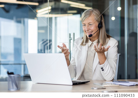 Senior smiling gray-haired woman in a suit and headset works in a call center, sits in the office at a laptop, consults and conducts an online video meeting, smiles and gestures with her hands. 111090433
