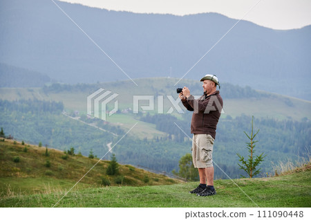 Male tourist hiking in hills, traveling in summer. Man standing on hill, holding camera, taking photo, admiring landscape, enjoying. Concept of tourism and adventure. Male tourist hiking in hills, traveling in summer. Man standing on hill, holding camera, taking photo, admiring landscape, enjoying. Concept of tourism and adventure. 111090448