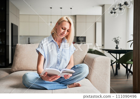 Woman sitting comfortably on couch, deeply engaged in reading book. Cozy living space with modern aesthetic on background. Person enjoying quiet moment of solitude, immersed in reading. 111090496