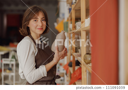 Smiling entrepreneur crafts woman holding mug in pottery studio while looking at camera Smiling entrepreneur crafts woman holding mug in pottery studio while looking at camera 111091039