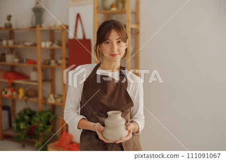 Portrait of young female potter in apron with mug looking at camera while posing in workshop Portrait of young female potter in apron with mug looking at camera while posing in workshop 111091067