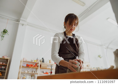 Smiling female potter in apron kneads piece of clay with her hands on table in studio Smiling female potter in apron kneads piece of clay with her hands on table in studio 111091097