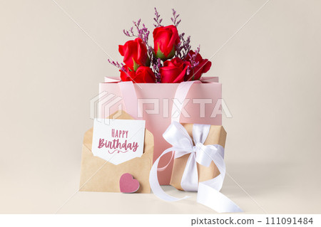 Happy birthday card and envelope with gift box and rose in paper bag on beige background Happy birthday card and envelope with gift box and rose in paper bag on beige background 111091484
