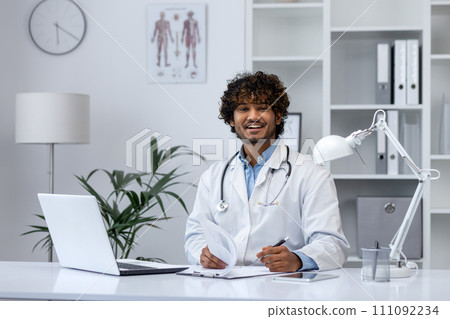 Confident young medical professional with stethoscope seated at a desk, using laptop in a bright clinic office. 111092234