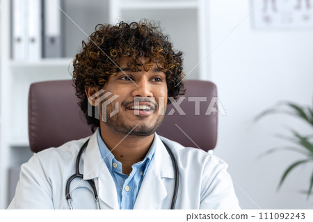 A young male Hindu doctor with curly hair smiling in a medical office, representing professionalism and care. 111092243
