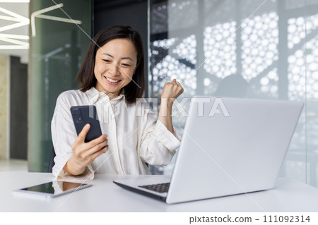 A cheerful young Asian businesswoman exudes success as she celebrates with a fist pump at her office workstation, laptop and phone in hand. A cheerful young Asian businesswoman exudes success as she celebrates with a fist pump at her office workstation, laptop and phone in hand. 111092314