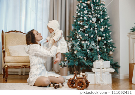 Brunette mother and little baby girl sitting near Christmas tree. Girls wearing a white dressing gowns. Woman holding her daughter in the air. 111092431