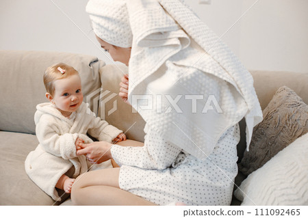 Young mother and little baby girl wearing dressing gowns with hair wrapped in towels. Woman and girl sitting on a sofa. Girl has a hairpins on her hair. 111092465