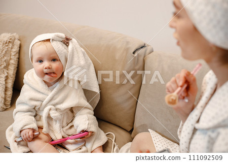 Focus on a little girl showing a tongue. Young mother and little baby girl wearing dressing gowns with hair wrapped in towels. Woman and girl sitting on a sofa. 111092509