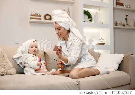 Young mother and little baby girl wearing dressing gowns with hair wrapped in towels. Woman and girl sitting on a sofa. Woman holding decorative cosmetics. 111092537