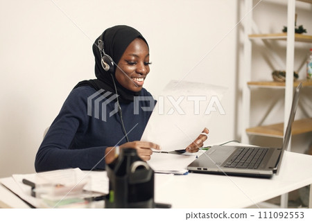 Young muslim business woman with laptop and file document sitting at table. Woman wearing blue dress and black hijab. Black woman using earphones. Young muslim business woman with laptop and file document sitting at table. Woman wearing blue dress and black hijab. Black woman using earphones. 111092553