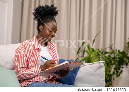 Woman writing her personal notes into her diary while sitting on sofa in living room. Woman writing her personal notes into her diary while sitting on sofa in living room. 111093853