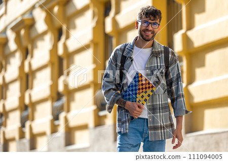 Male student in glasses holding textbooks and smiling while standing at university building. Male student in glasses holding textbooks and smiling while standing at university building. 111094005