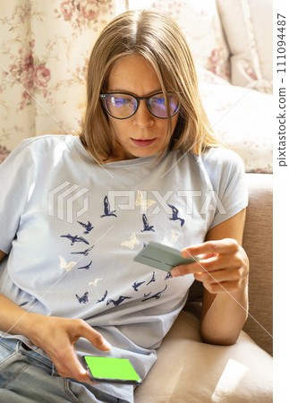 Young woman sitting at home with smartphone with green screen and bank card in hands. Online payment 111094487
