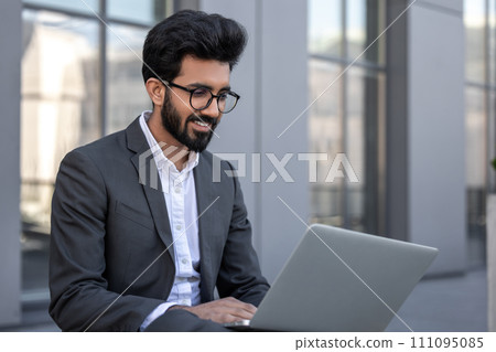 Young hindu businessman working on a laptop while sitting near the office building 111095085