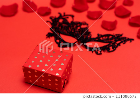 Valentine's day black lace mask andgiftbox with gold hearts on red background with rose petals 111096572