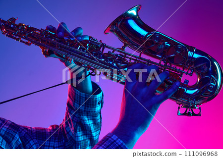 Cropped portrait of jazzman playing saxophone against gradient blue-pink background in neon light. Jazz melodies. Cropped portrait of jazzman playing saxophone against gradient blue-pink background in neon light. Jazz melodies. 111096968