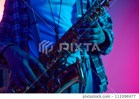 Cropped portrait of jazzman holds saxophone against gradient blue-pink background in neon light. Jazz melodies. Cropped portrait of jazzman holds saxophone against gradient blue-pink background in neon light. Jazz melodies. 111096977