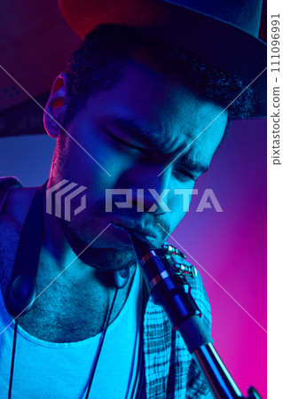 Close up portrait of young man, focused artist in retro hat playing saxophone against gradient studio background in mixed neon light. Close up portrait of young man, focused artist in retro hat playing saxophone against gradient studio background in mixed neon light. 111096991