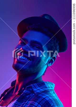 Portrait of African-American artist, Jazzman in hat smiling looking away in neon light against gradient blue-pink background. 111096996