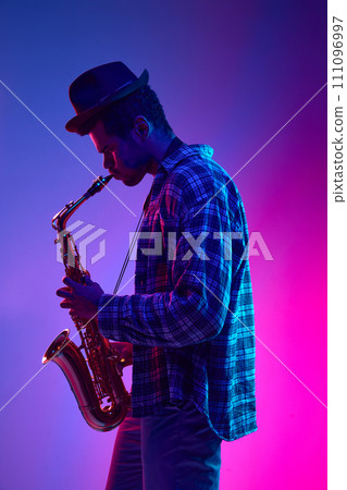 Portrait of talented solo saxophonist playing piece against pink-blue gradient background in neon light. Ad 111096997