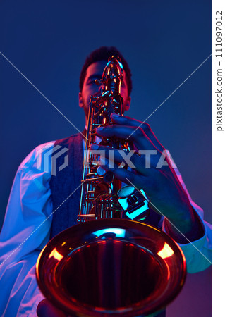 Close up portrait. African-American talented man, musician playing saxophone against studio background in blue and pink lighting. Close up portrait. African-American talented man, musician playing saxophone against studio background in blue and pink lighting. 111097012