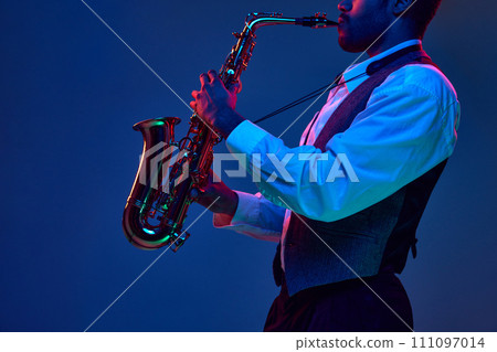 African-American jazz musician playing saxophone his major melodies against blue background in mixed pink-purple lighting. 111097014