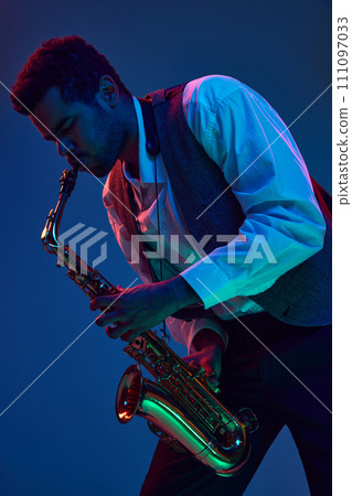 African-American man virtuoso, solo musician playing jazz melodies on saxophone against gradient blue background in mixed neon light. 111097033