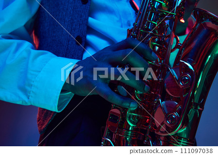 African-American artist, solo performer plays chords on fingering of saxophone, wind instrument in neon light against studio background. African-American artist, solo performer plays chords on fingering of saxophone, wind instrument in neon light against studio background. 111097038