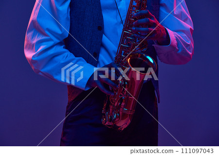 Cropped portrait of young man, saxophonist, musician in shirt and vest holds wind instrument against studio background in neon lighting. 111097043