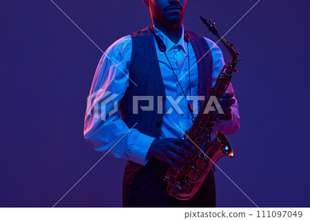 Portrait of young man, jazz performer, instrumental musician holds saxophone and looking at camera against dark background in neon light. Portrait of young man, jazz performer, instrumental musician holds saxophone and looking at camera against dark background in neon light. 111097049