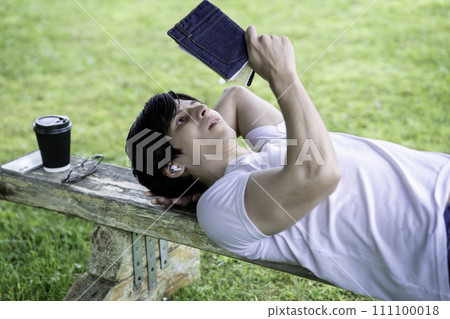 A foreign man lying on a park bench in fresh greenery, listening to music and reading a book. A foreign man lying on a park bench in fresh greenery, listening to music and reading a book. 111100018